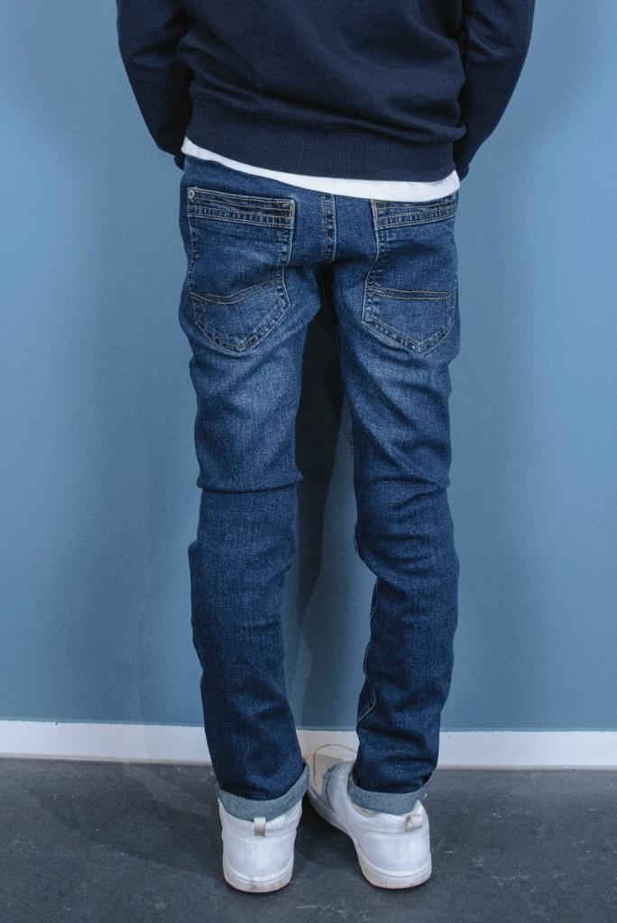 Boy wearing blue jeans and white shoes against a light blue wall.