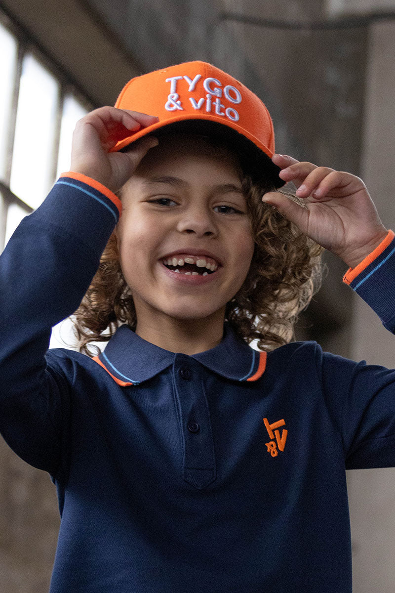 A smiling child wearing a navy long-sleeve polo shirt with orange and white trim, an orange cap with &