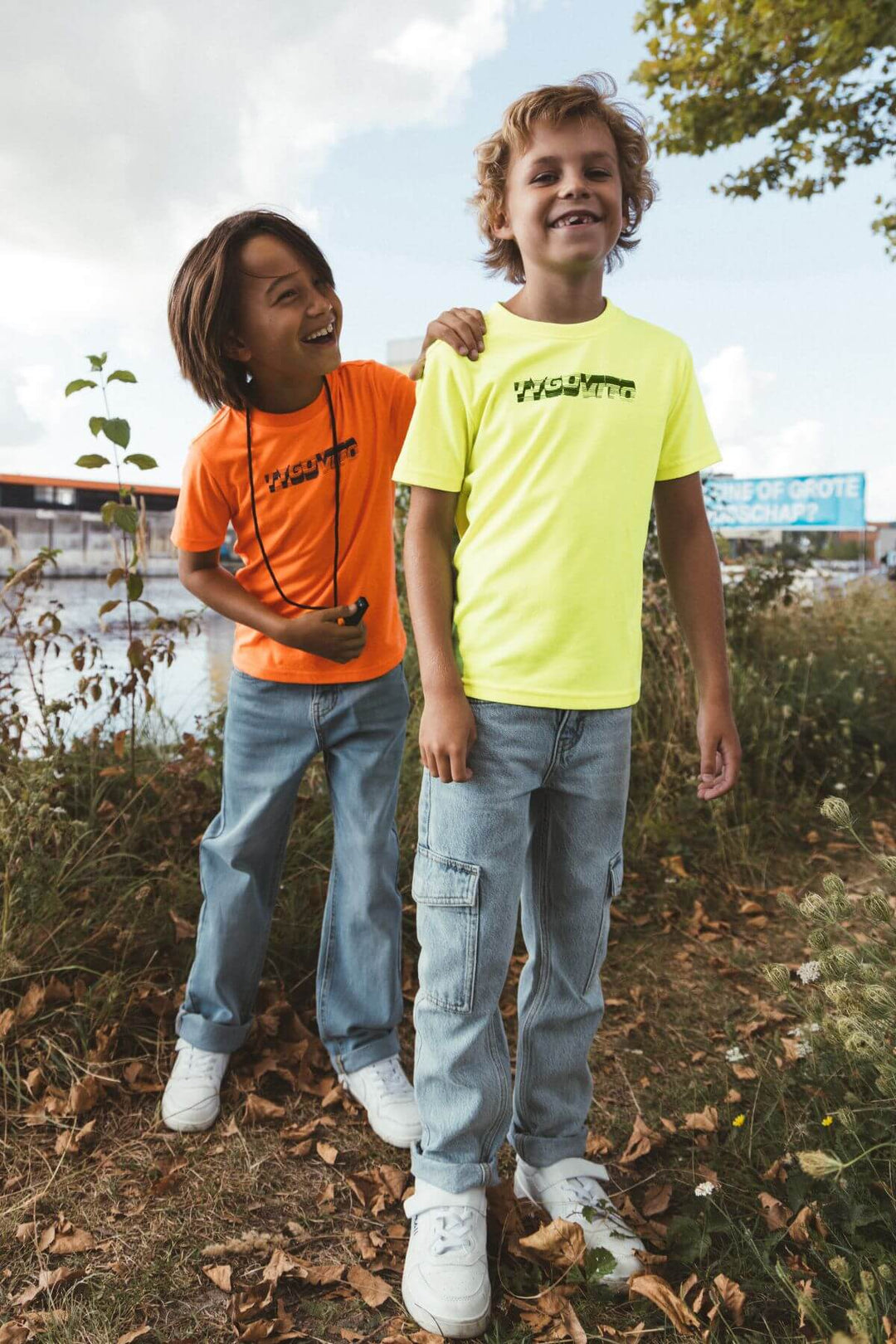 Two children standing outdoors with a lake and trees in the background