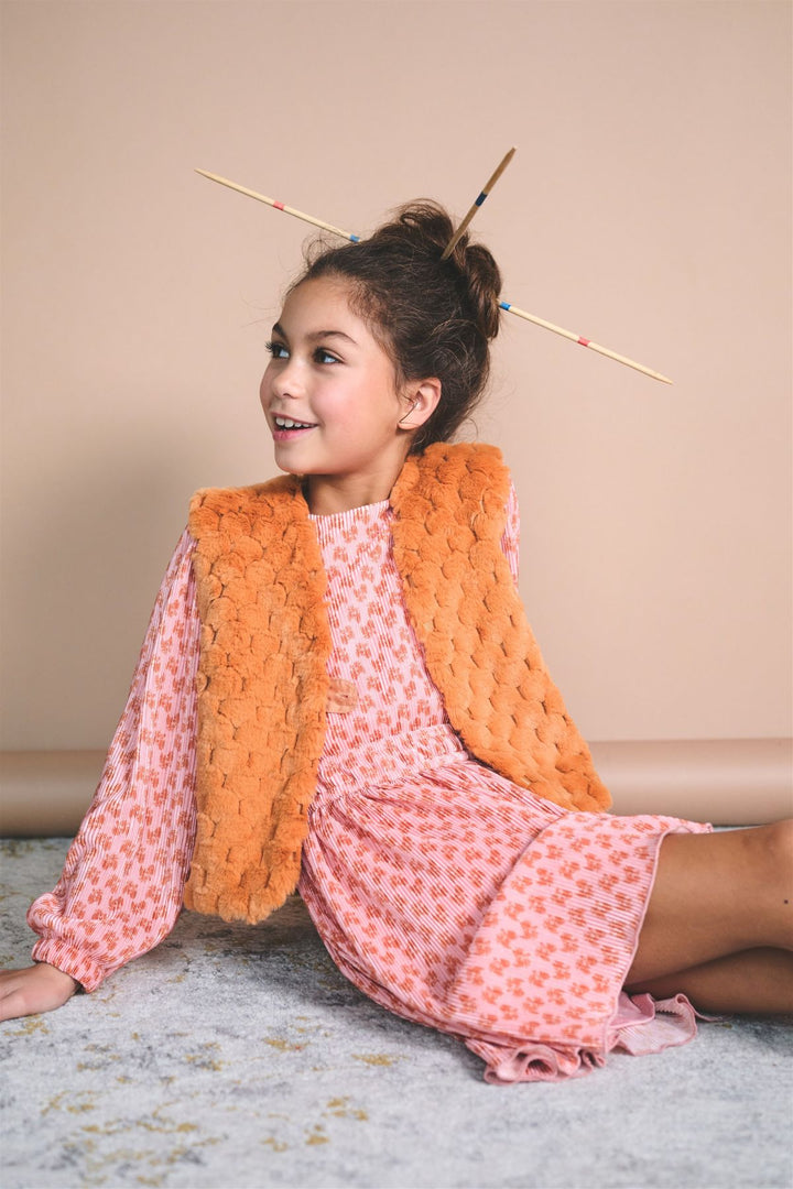 A young girl sits on a light gray rug against a beige wall, wearing a pink patterned dress and a chunky orange textured vest. She has wooden hair sticks in her hair.