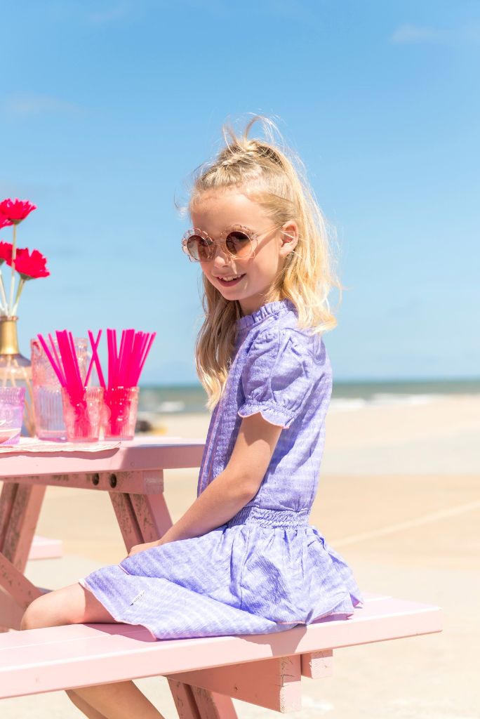 Young girl in the purple dress Dotty sitting at a pink table on a beach.