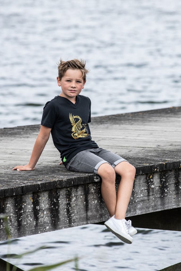 Boy sitting down by the water and wearing the grey denim shorts with a black T-Shirt